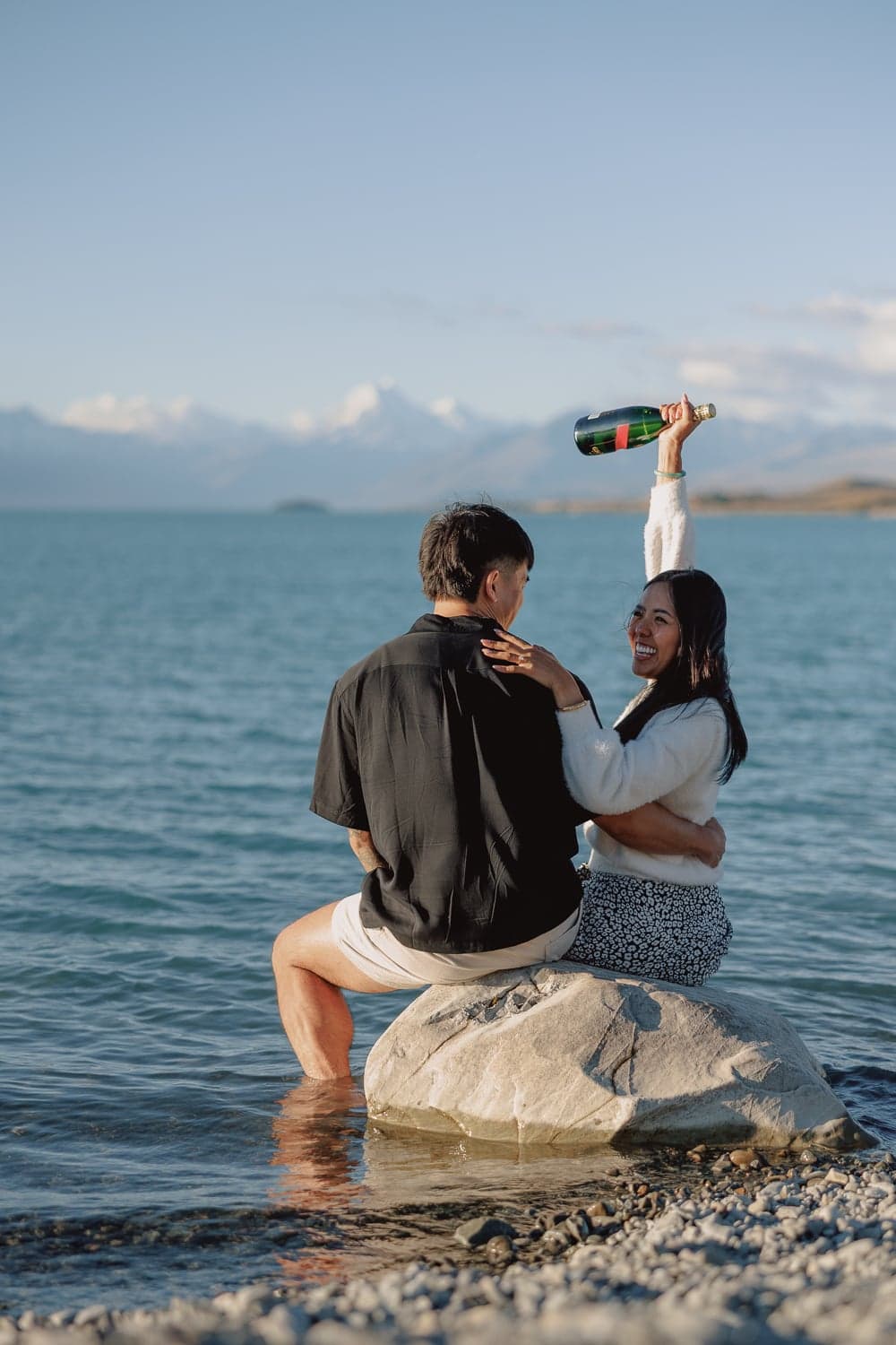 Newly engaged woman raises champagne bottle on rocks during proposal photography at Lake Pukaki, New Zealand.