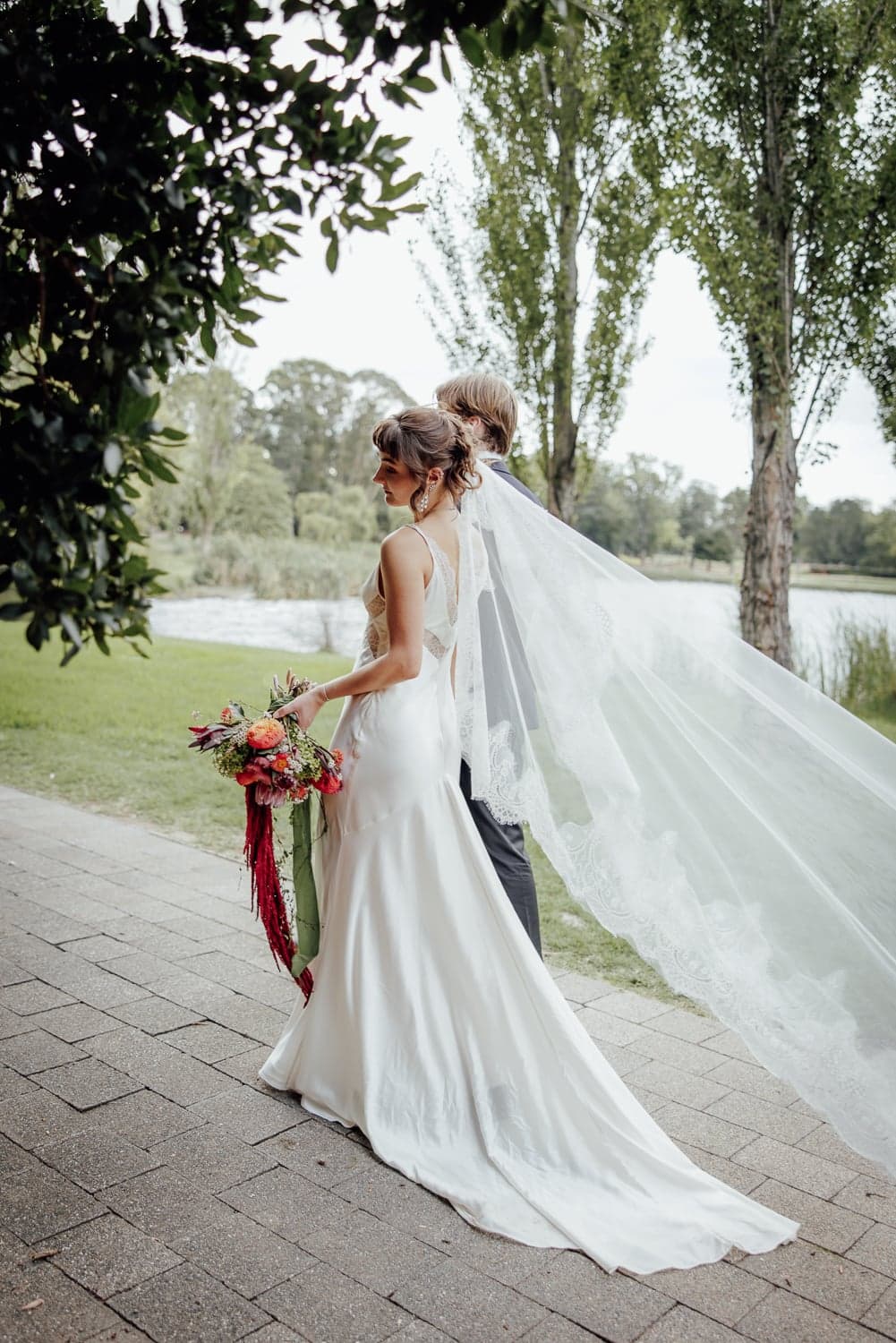 Bride and groom walking along the lakeside path at Commonwealth Park, Canberra, with the bridal veil trailing in the wind.