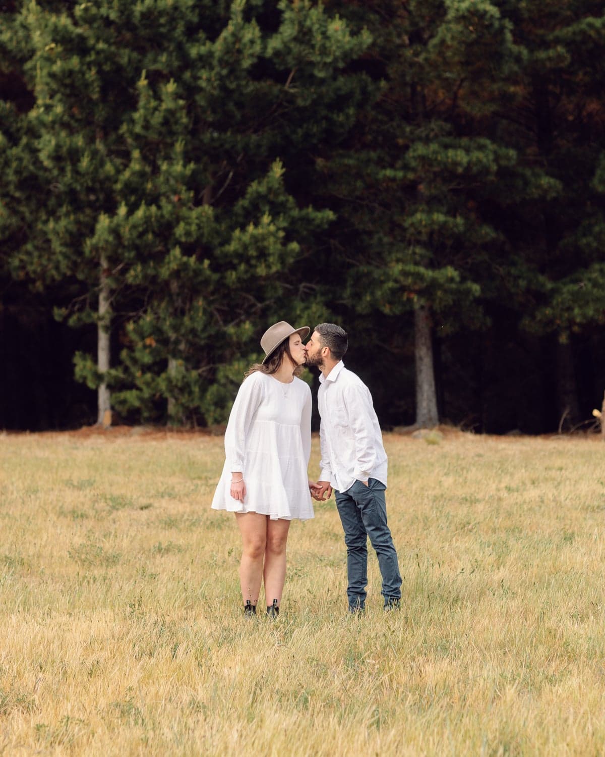 Couple kissing in a golden grass field backed by pine trees during a Canberra engagement shoot.
